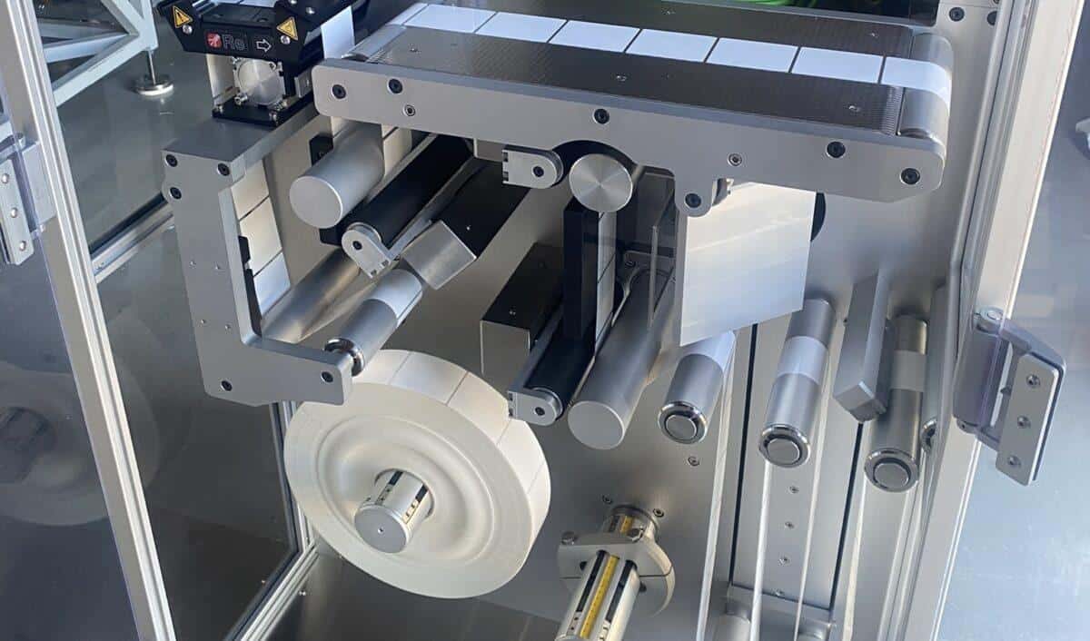 Close-up of the PharmaJet label web path: liner roll, rollers, peel plate and product transport belt inside the machine